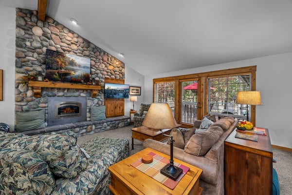 Unwind in your spacious living room featuring a stunning stone fireplace, vaulted ceilings, and natural light streaming through large windows overlooking the forest.
