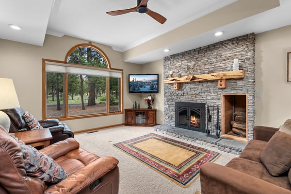 Sink into plush leather sofas before your stacked-stone fireplace, where a ceiling fan whispers overhead and picture windows frame serene woodland views—your cozy retreat awaits.