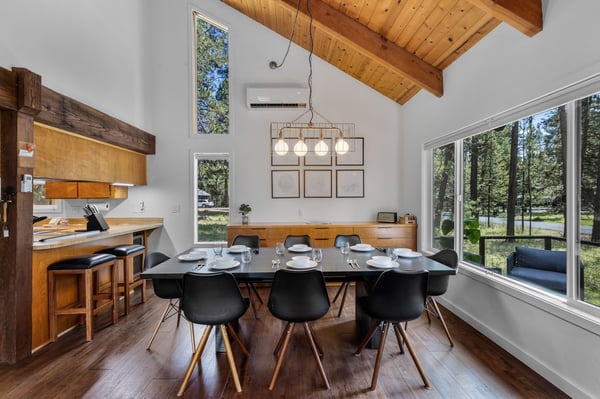 Gather around your spacious dining table, where natural light pours through towering windows framing forest views. Warm wood beams overhead and an open kitchen nearby create a cozy, inviting space perfect for memorable meals together.