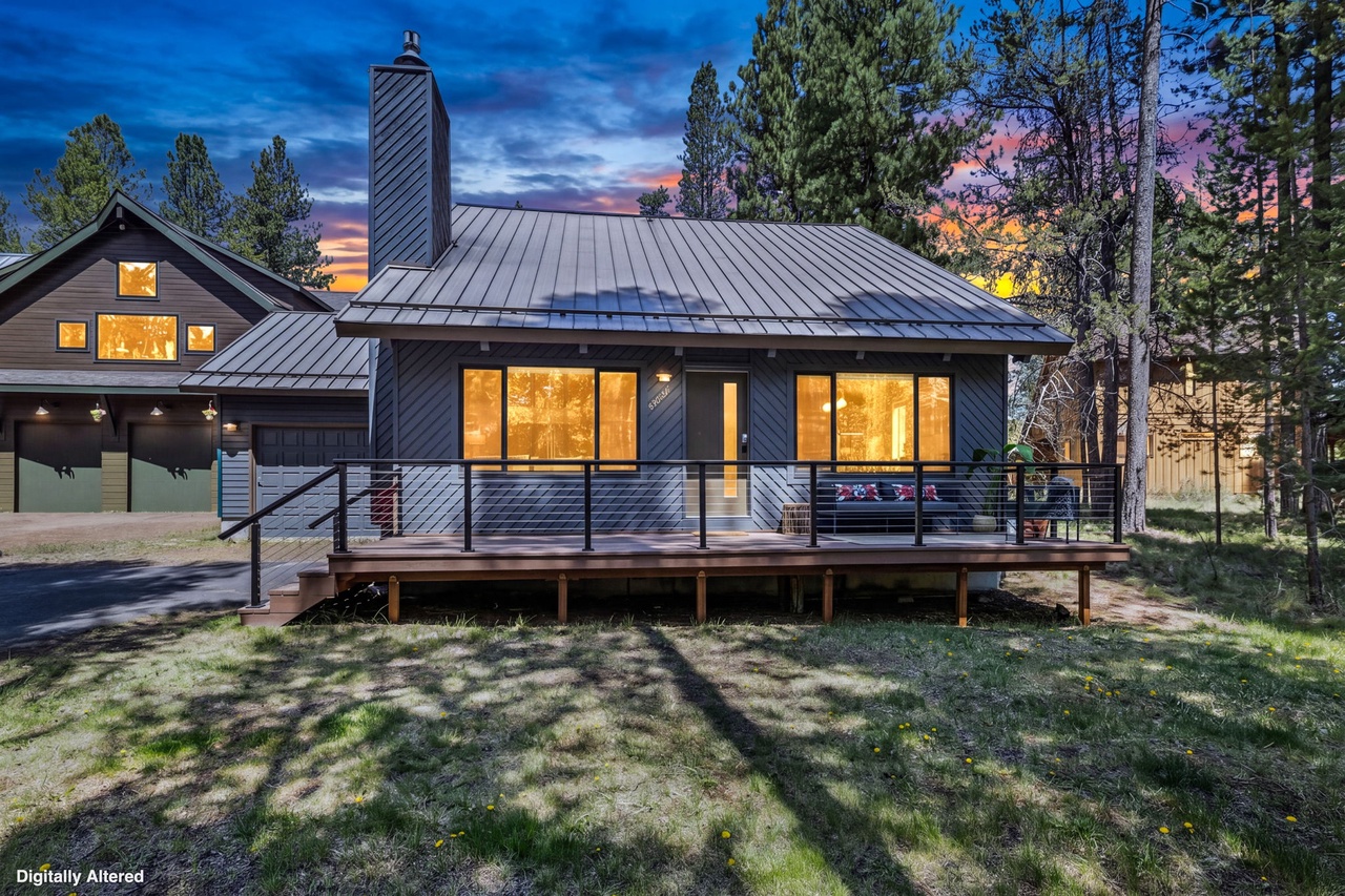 Modern mountain retreat surrounded by towering pines with a striking sunset backdrop. The property's sleek dark siding and warm interior lighting create a welcoming contrast to the natural landscape.