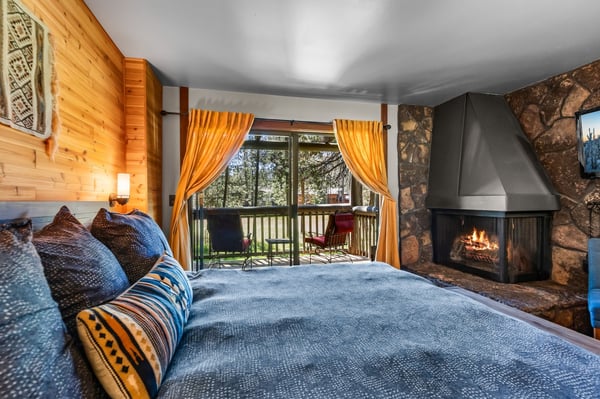 Unwind in this cozy bedroom where warm wood tones meet rustic stone, a crackling fireplace sets the mood, and your private balcony overlooks the peaceful forest.